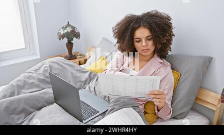 Femme hispanique aux cheveux bouclés examine des documents dans une chambre avec un ordinateur portable à proximité, transmettant un sentiment de travail à domicile. Banque D'Images