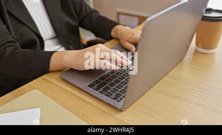 Femme hispanique avec des cheveux bouclés travaillant sur ordinateur portable dans un cadre de bureau présente un jeune adulte attrayant dans une tenue professionnelle à l'intérieur. Banque D'Images