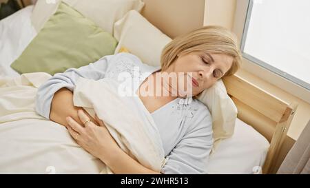 Une femme blonde sereine d'âge moyen dormant paisiblement seule dans une chambre ensoleillée, exsudant la tranquillité. Banque D'Images