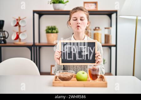 Femme blonde comique faisant visage de poisson fou tout en profitant d'un petit déjeuner sain à la maison, tableau noir à la main Banque D'Images