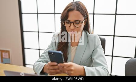 Séduisante jeune femme hispanique, sérieuse mais détendue, patron dans son bureau, axée sur le succès, textant professionnellement sur son smartphone, tout en elegantl Banque D'Images