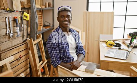 Un homme afro-américain sourit assis en toute confiance dans son atelier de menuiserie rempli de divers outils et équipements. Banque D'Images