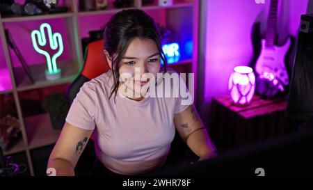 Femme hispanique souriant dans une salle de jeux éclairée au néon la nuit avec une toile de fond d'une guitare électrique et de lumières décoratives. Banque D'Images