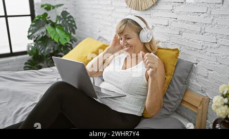 Femme blonde d'âge moyen confiante, enveloppée dans son pyjama, assise détendue sur son lit, interagissant avec un sourire sur un vide matinal chaud et étincelant Banque D'Images