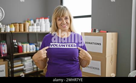 Femme blonde d'âge moyen, pleine de joie, pointe vers son uniforme de bénévole tout en rayonnant un sourire confiant au centre caritatif animé, sentant l'unité Banque D'Images