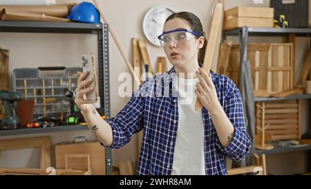 Une jeune femme caucasienne portant des lunettes de sécurité participe à un appel vidéo dans son atelier de menuiserie à l'intérieur. Banque D'Images