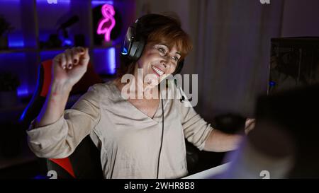 Une femme mature joyeuse avec un casque célèbre dans une salle de jeu éclairée au néon la nuit Banque D'Images