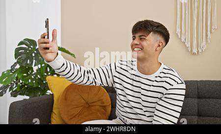 Joyeux jeune homme hispanique capturant joyeusement un selfie avec smartphone tout en se refroidissant sur le canapé dans le salon confortable de la maison. Banque D'Images