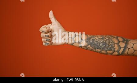 Main d'homme avec le pouce tatoué donnant un geste d'approbation du pouce vers le haut sur un fond rouge isolé - un signe positif d'un mâle hispanique adulte! Banque D'Images