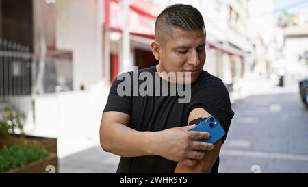 Jeune homme latin confiant, heureux scannant son capteur de diabète dans la rue de la ville à l'aide de son smartphone, surveillant de manière experte son taux de glucose, putti Banque D'Images