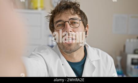 Bel homme barbu avec des lunettes en manteau blanc posant à l'intérieur d'une clinique dans un portrait de style selfie. Banque D'Images