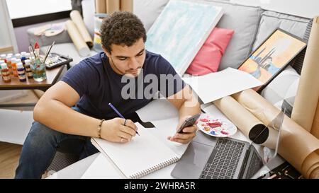 Jeune homme dessinant dans un studio d'art tout en vérifiant son smartphone, entouré de peintures et de fournitures d'art. Banque D'Images