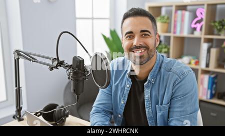 Un homme hispanique souriant avec une barbe dans un cadre de studio avec microphone et ordinateur portable, projetant un comportement professionnel mais décontracté. Banque D'Images