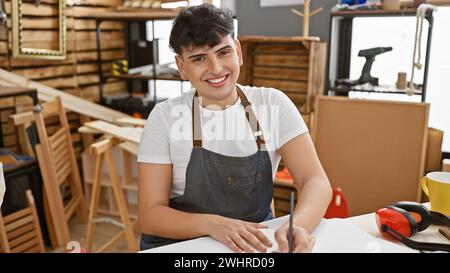 Homme souriant portant un tablier prenant des notes dans un atelier de menuiserie créative, entouré de matériaux et d'outils en bois. Banque D'Images