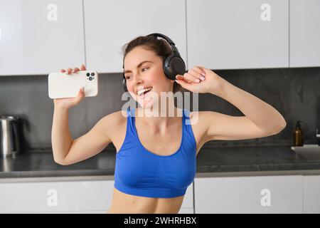 Portrait de belle blogueuse de fitness, femme au casque, écoutant de la musique et dansant dans la cuisine, portant des leggings bleus et sp Banque D'Images