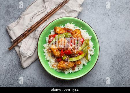 Poulet collant aigre-doux asiatique avec légumes sautés et riz dans un bol en céramique avec baguettes vue de dessus, ston rustique gris Banque D'Images