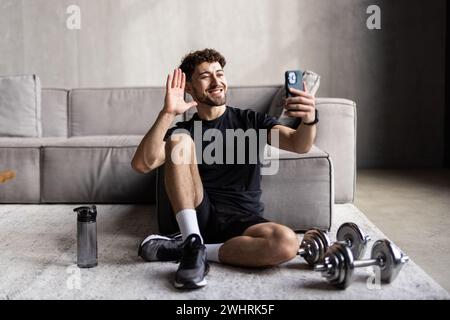 Souriant millénaire un homme européen musclé assis sur le sol en position lotus, en agitant la main à l'ordinateur dans le salon intérieur.Sport style de vie, blog A. Banque D'Images