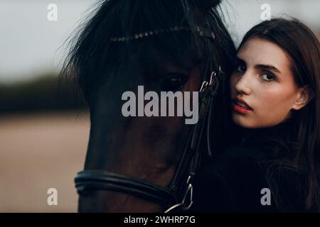 Jeune femme en noir avec son cheval au coucher du soleil dehors. Banque D'Images