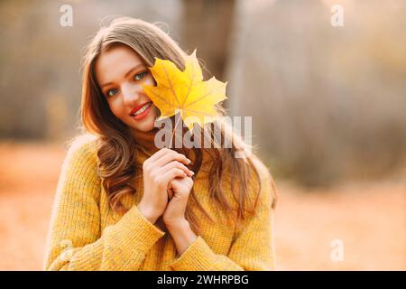 Jeune modèle de femme dans le parc d'automne tient dans sa main feuille d'érable jaune feuillage au visage Banque D'Images