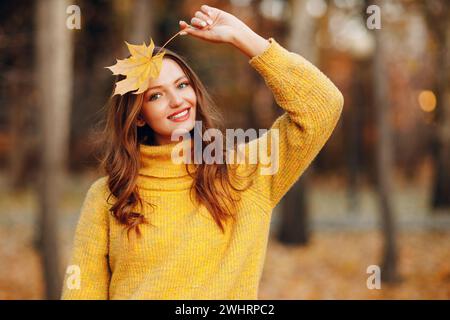 Jeune modèle de femme dans le parc d'automne tient dans sa main feuille d'érable jaune feuillage au visage Banque D'Images