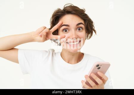 Portrait de la jeune femme montre le signe de la paix, tient un smartphone, écoute de la musique dans des écouteurs sans fil, se tient au-dessus du backgroun blanc Banque D'Images