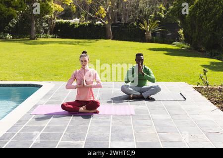 Couple diversifié pratiquant le yoga et méditant à côté de la piscine dans le jardin Banque D'Images