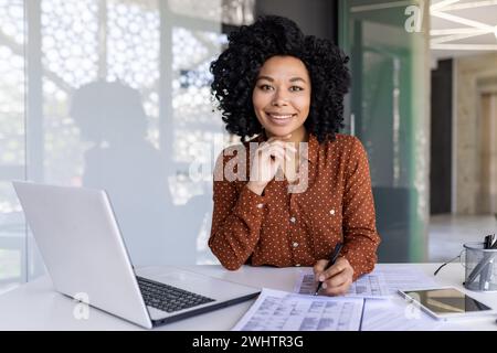 Femme latino en blouse à pois remplissant les tables à la main tout en travaillant au bureau techniquement équipé. Administrateur de bureau motivé passant commande pour fournir à l'entreprise les fournitures nécessaires. Banque D'Images