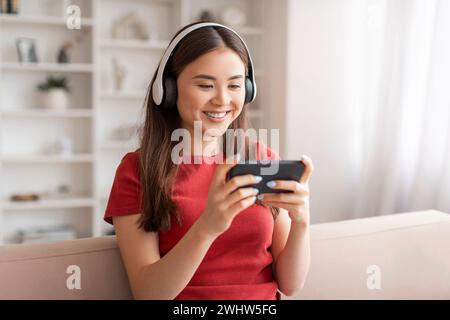 Jeune femme asiatique souriante portant des écouteurs s'engageant avec son smartphone à la maison Banque D'Images