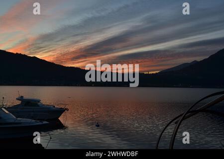 Beau coucher de soleil rouge avec des bateaux à moteur ancrés à la jetée du lac et un détail de bateau de pêche en bois traditionnel italien du lac de Côme 'Lucia'. Grand ciel sombre. Banque D'Images