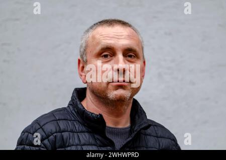 Un homme âgé aux cheveux gris de 45-50 ans, contre un mur de couleur claire portant une veste noire, regarde la caméra. Banque D'Images