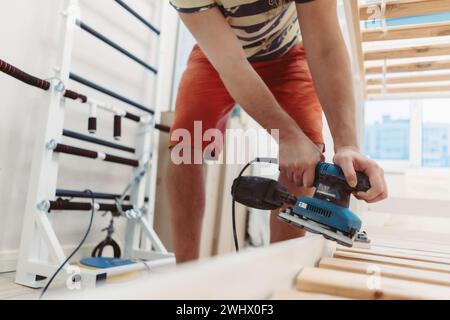 l'homme méconnaissable frotte le lit d'enfant en bois avec une ponceuse électrique ou un broyeur en enlevant la vieille peinture Banque D'Images