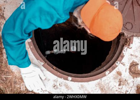 Un plombier se pencha au-dessus du puits pour vérifier le compteur d'eau. Travaux de plomberie en milieu rural. Banque D'Images