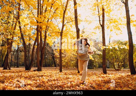 Jeune femme souriante joue et saute dansant dans le parc d'automne avec les feuilles jaunes au coucher du soleil. Banque D'Images