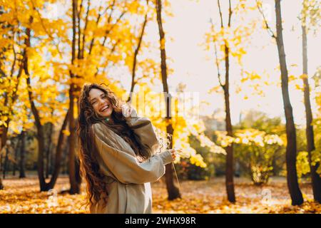 Jeune femme souriante joue et saute dansant dans le parc d'automne avec les feuilles jaunes au coucher du soleil. Banque D'Images