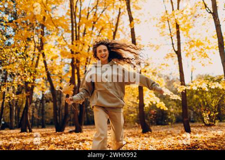 Jeune femme souriante joue et saute dansant dans la forêt d'automne avec les feuilles jaunes au coucher du soleil. Banque D'Images