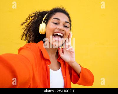 Perspective selfie d'un adolescent afro-américain avec des écouteurs s'amusant avec un mur jaune en arrière-plan Banque D'Images