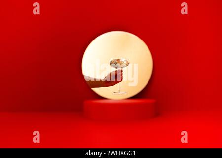 Miroir rond avec une scène sur un fond rouge vif. Main féminine blanche avec une manucure rouge tenant un verre de champag pétillant Banque D'Images