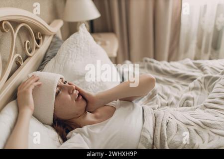 Jeune femme avec mal de tête grippe malade maladie froide à la maison à l'intérieur couché sur le lit avec compresse froide sur sa tête. Banque D'Images