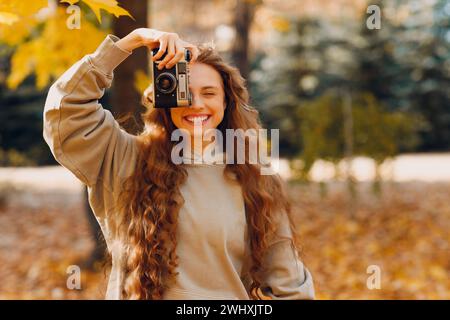 Jeune femme souriante avec la caméra photo photographiant les arbres de la forêt avec les feuilles jaunes au coucher du soleil. Banque D'Images