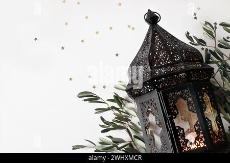 Brindilles d'olivier marocain foncé éclatant, lanterne arabe et rameaux d'olivier isolées sur fond de table blanc. Confettis étoiles dorées. Nature morte festive, bannière Banque D'Images