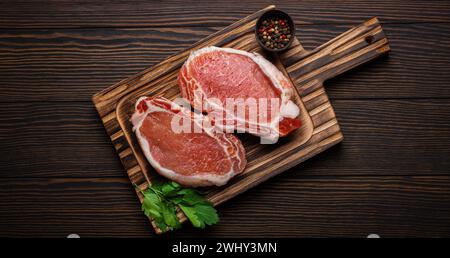 Couper les steaks de porc crus avec des assaisonnements sur la planche à découper de cuisine, fond en bois rustique d'en haut, prêt pour le barbecue. Longe de porc Banque D'Images