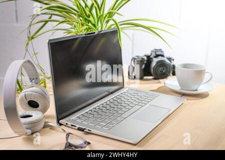 Ordinateur portable, écouteurs et appareil photo sur un bureau en bois avec tasse à café une plante contre un mur blanc, affaires créatives, copie spac Banque D'Images