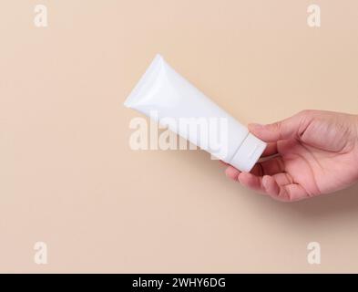 La main d'une femme tient un tube en plastique blanc pour crème, dentifrice sur fond beige Banque D'Images