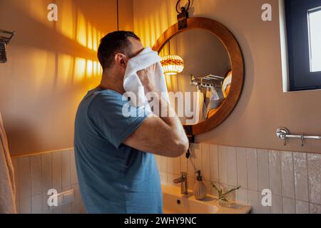 Jeune homme attrayant regarde par la fenêtre et s'essuie les mains avec une serviette blanche dans sa salle de bain. Un homme caucasien se lave le visage an Banque D'Images