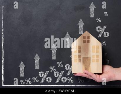 Une main tient une maison en bois sur le fond d'un graphique avec des indicateurs croissants, le concept de la hausse des prix de l'immobilier Banque D'Images