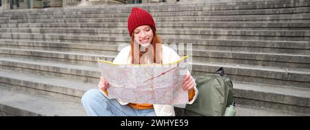 Portrait d'une jeune femme à tête rouge, le touriste se trouve avec une carte papier et cherche un itinéraire vers l'attraction touristique, repose sur les escaliers outdoo Banque D'Images