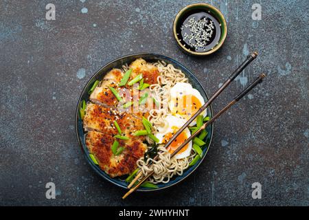 Nouilles asiatiques soupe ramen avec filet de poulet panko frit et œufs durs dans un bol en céramique avec bâtonnets hachés, sauce soja sur sto Banque D'Images