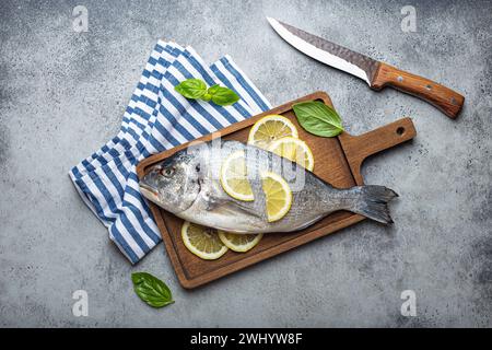 Poisson cru dorado en cocotte avec ingrédients citron, basilic frais, bouteille d'huile d'olive sur planche à découper en bois avec couteau sur Banque D'Images