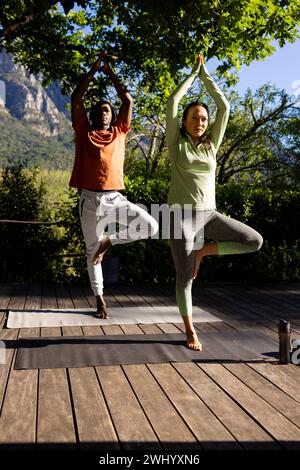 Heureux couple diversifié pratiquant le yoga debout sur le pont dans le jardin ensoleillé, espace de copie Banque D'Images
