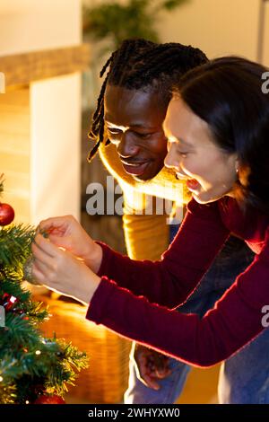 Heureux couple diversifié ayant du plaisir à décorer l'arbre de noël à la maison Banque D'Images